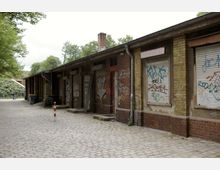 Backstein-Gebäude mit verschlossenen, mit Graffiti bemalten Rolläden, umgeben von Kopfsteinpflaster und Bäumen im Hintergrund. Ein einzelner rot-weißer Poller steht in der Mitte des Weges vor dem Gebäude.