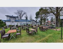 Außenbereich eines Restaurants mit großen Holztischen und geflochtenen Stühlen auf einer Rasenfläche. Im Hintergrund ein weißes Gebäude mit blauen Dächern, Glasfassade und umgeben von hohen Bäumen.