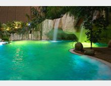 Innenansicht eines tropischen Schwimmbades mit einem Wasserfall, der von einer Felswand in ein türkisfarbenes Becken fällt. Im Hintergrund sind üppige Vegetation mit Palmen und Kletterpflanzen sowie eine Kuppelstruktur zu sehen.