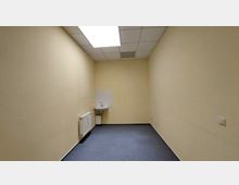 Kleiner, leerer Raum mit cremefarbenen Wänden, blauer Auslegware und einer Deckenleuchte. An der Stirnwand befindet sich ein kleines Waschbecken neben einem Heizkörper.