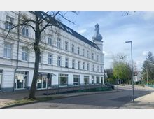 Historisches weißes Gebäude mit drei Etagen, großen Fenstern und einem markanten Zwiebelturm auf dem Dach, umgeben von Bäumen und einer gepflasterten Straße. Im Vordergrund ein Einzelbaum, Gehweg und Rampen, die zum Eingang führen.