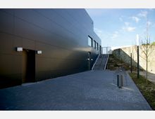 Moderne Außenansicht eines Gebäudes mit glatter, dunkelgrauer Fassade, einem Eingang links sowie einem gepflasterten Vorplatz. Rechts neben dem Gebäude führen Treppenstufen zu einem höher gelegenen Bereich, begleitet von einem Geländer; daneben befinden sich kleine Bäume und ein steinernes Mauerelement.