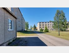 Mehrstöckige Plattenbauten in einem Wohngebiet, umgeben von Rasenflächen mit vereinzelt stehenden Bäumen. Ein Gehweg führt zwischen den Gebäuden hindurch, begleitet von einer Straßenlaterne.