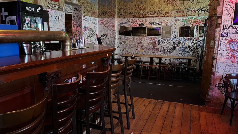 Innenraum einer kleinen Bar mit langer Holztheke und hohen Barhockern auf Holzdielenboden. Die Wände sind dicht mit Graffiti und Kritzeleien bedeckt, daneben stehen schmale Tische mit Stühlen und eine verspiegelte Wand mit Lampen.