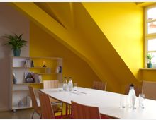 Ein moderner Besprechungsraum mit einer weißen Tischplatte, umgeben von Holzstühlen, vor einer schräge gelben Wand mit einem Fenster. Ein Regal an der linken Wand enthält Bücher, Dekorationsobjekte und eine Pflanze in einem grünen Topf.