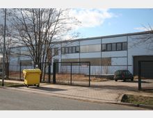 Industrielle Lagerhalle mit hellblauer Fassade und großen Fenstern, umgeben von einem Metallzaun. Im Vordergrund ein gelber Müllcontainer und ein parkendes Auto; der Eingangsbereich liegt auf einem gepflasterten Weg.