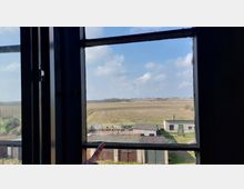 Blick aus einem Fenster auf eine ländliche Szenerie mit weiten Feldern, kleinen Gebäuden und einem hellen Himmel mit Wolken. Im Vordergrund ist ein Rahmen des Fensters und eine Hand sichtbar.