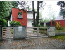 Modernes Wohnhaus mit roten und weißen Fassaden, umgeben von Bäumen und einem Garten. Vor dem Haus befindet sich ein Metallzaun mit einem Eingangstor aus Beton und Holzlatten.