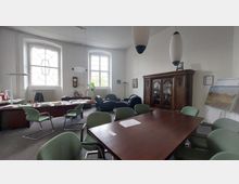 Ein Büro- und Konferenzraum mit einer großen Holztischgruppe und grünen Stühlen, im Hintergrund ein Schreibtischbereich mit großen Fenstern. Ein dunkler Holzschrank und ein Gemälde auf einem Ständer sind an der Wand zu sehen, während moderne Hängelampen von der Decke hängen.