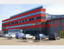 Modernes Bürogebäude mit einer Fassade aus roten und schwarzen horizontalen Streifen und großen Fenstern, davor ein unbefestigter Parkplatz mit geparkten Autos und einem Bus.