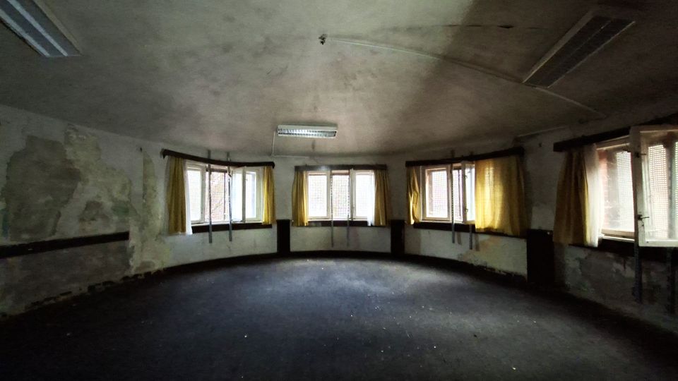 Leerer, runder Innenraum eines verlassen wirkenden Gebäudes mit abgenutzten Wänden, dunkelgrauem Boden und einer flachen Decke mit Leuchtstofflampen. Entlang der gebogenen Außenwand befinden sich mehrere Fenster mit teils gelben Vorhängen, durch die Tageslicht einfällt.