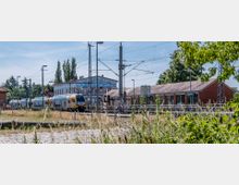 Bahnhofsbereich mit einem Regionalzug, der auf einem Gleis neben einem roten Backsteingebäude steht. Im Hintergrund sind ein bemaltes Gebäude, Oberleitungen und Bäume zu sehen, während im Vordergrund Gräser und Sträucher wachsen.