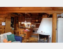 Gemütlicher Innenraum eines Blockhauses mit Holzwänden und -balken. Zu sehen ist eine offene Küche mit Holzschränken, ein Esstisch mit Stühlen sowie ein heller, gemauerter Kamin im Vordergrund.