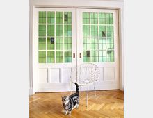 Eine helle Inneneinrichtung mit weißen Schiebetüren, die grüne Glasfenster mit kleinen farbigen Wappenmotiven zeigen. Im Vordergrund stehen ein weißer Metallstuhl und eine getigerte Katze auf einem Parkettboden.
