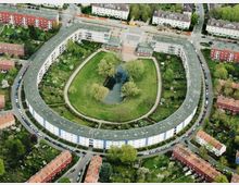 Luftaufnahme eines halbkreisförmigen Wohngebäudekomplexes, der eine grüne Anlage mit einem kleinen Teich in der Mitte umschließt. Die Umgebung ist von weiteren Wohnhäusern, Gärten und Straßen geprägt.