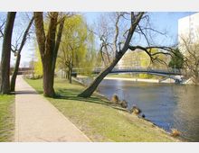 Grüne Parklandschaft mit einem gepflasterten Weg, der entlang eines Flusses verläuft. Im Hintergrund führt eine moderne Fußgängerbrücke über das Wasser, umgeben von Bäumen, darunter eine Trauerweide mit hängenden Ästen.