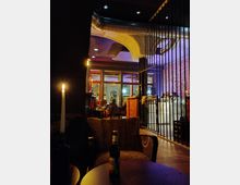 Innenraum eines gemütlich beleuchteten Cafés mit Sitzbereichen aus Polstermöbeln und einem runden Holztisch, auf dem eine brennende Kerze und eine Bierflasche stehen. Im Hintergrund sind eine Bar, Gitterelemente, sichtbare Lüftungsrohre an der Decke und große Fenster sichtbar.