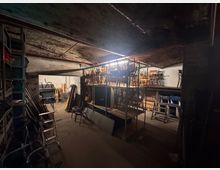 Ein Kellerraum mit gewölbter Decke und rohen Betonwänden, der als Lager genutzt wird. Verschiedene gestapelte Stühle, Leitern und Metallregale sind sichtbar, teilweise auf erhöhten Plattformen gelagert.