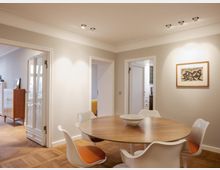 Essbereich in einem modernen Wohnraum mit einem runden Holztisch, weißen Stühlen mit orangefarbenen Sitzpolstern und einer Schale als Tischdekoration. Der Raum hat hell gestrichene Wände, Parkettboden, eine Kunstzeichnung an der Wand und offene Türen, die zu weiteren Räumen führen.