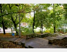 Ein kleiner Parkbereich mit gepflasterten Wegen, einer niedrigen Steinmauer, vielen Bäumen und Laub auf dem Boden. Im Hintergrund ist ein Gebäude mit einem Fenster sichtbar, hinter einer von Bäumen gesäumten Straße.