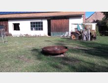 Ein ländlicher Garten mit einer Feuerstelle im Vordergrund, einem weißen Gebäude mit Holzdetails und Solarpaneelen auf dem Dach im Hintergrund. Rechts steht ein alter Traktor neben einem Stapel Holz, daneben sind Gartenmöbel erkennbar.