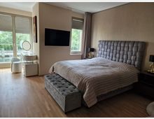 Modernes Schlafzimmer mit einem großen Bett und gestepptem grauem Kopfteil, flankiert von Nachttischen mit Lampen. Links befindet sich ein Schminktisch mit Hocker vor einer Fensterfront, die auf einen Balkon mit Glasgeländer führt, und rechts ein weiteres Fenster mit Blick auf grüne Bäume.