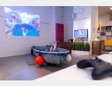 Ein modern gestalteter Ausstellungsraum mit einem kleinen blauen Ruderboot in der Mitte, dekoriert mit Rettungsringen und Bojen. Im Hintergrund sind eine Wandprojektion mit einer spielähnlichen Wasserlandschaft, ein Bildschirm neben Pflanzen und Holzmöbeln sowie weitere Exponate zu sehen.