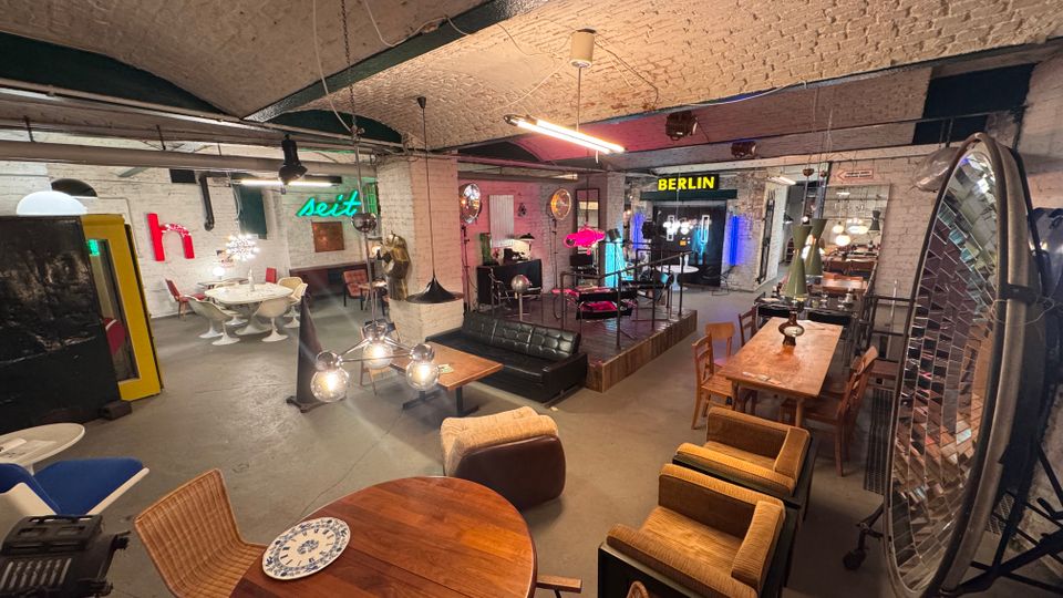 Ein loftartiger Raum mit gewölbter Ziegeldecke, eingerichtet mit unterschiedlichen Retro-Möbeln, Tischen und Stühlen. Im Hintergrund sind Neonlichter, Schilder mit "BERLIN" und "seit", sowie moderne Lampen und Dekorationen sichtbar.
