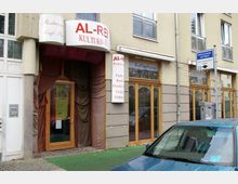 Außenansicht eines Gebäudes mit dem Schild "AL-REI Kulturhaus" über dem Eingang, der mit verspielten Stoffvorhängen dekoriert ist. Rechts daneben befinden sich große Fenster und ein Schild mit der Aufschrift "Kaffeehaus"; ein Parkplatz mit einem blauen Auto liegt im Vordergrund.