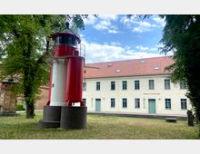 Ein kleiner roter Leuchtturm steht in einem begrünten Außenbereich vor dem Gebäude des Museums Fürstenwalde mit cremefarbener Fassade, grünen Fensterrahmen und einem roten Ziegeldach. Links neben dem Leuchtturm sind ein großer Baum und ein historisches Objekt zu sehen.