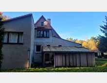 Haus im ländlichen Stil mit verputzter Fassade und Satteldach. Im Vordergrund befindet sich ein Anbau mit einem verglasten, teilweise mit Vorhängen verdeckten Wintergarten, umgeben von Grünfläche und Bäumen im Hintergrund.