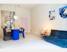 Minimalistisch eingerichteter Wohnraum mit hellen Wänden. Im Vordergrund ein blauer, gepolsterter Sessel, ein runder Marmortisch mit Büchern und einer roten Tischlampe, dahinter ein runder blauer Esstisch mit schwarzen und blauen Stühlen. An der Wand sind moderne abstrakte Gemälde und eine große Glaswand erkennbar.