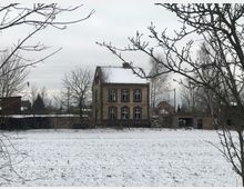 Ein altes, zweistöckiges Gebäude mit zerbrochenen Fensterscheiben und einer schneebedeckten Dachfläche steht inmitten einer offenen, verschneiten Wiese. Im Hintergrund sind kahle Bäume, Nebengebäude und ein trüber Himmel zu sehen.