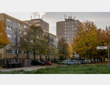Mehrstöckige Wohngebäude aus Beton in einer Wohnsiedlung, umgeben von herbstlich gefärbten Bäumen. Im Vordergrund stehen geparkte Autos, und ein Straßenschild mit der Aufschrift „Mühlenweg“ ist sichtbar.