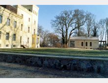 Gelbes klassizistisches Gebäude mit einem Turm auf der linken Seite, daneben ein kleineres Nebengebäude mit Rundbogenfenstern. Im Vordergrund befindet sich eine Rasenfläche, begrenzt durch eine niedrige Steinmauer, und im Hintergrund stehen große, unbelaubte Bäume.