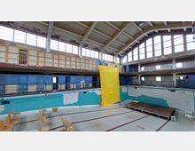 Ein leerstehendes Hallenbad mit hoher Decke und vielen Fenstern. Das Becken ist trocken, an den Wänden sind blaue und grüne Fliesen, und ein großes gelbes Tuch hängt von der oberen Etage herab; vereinzelte Stühle und eine kleine Bühne stehen im Becken.