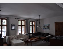 Wohnzimmer mit großen Holzrahmenfenstern, die viel Licht hereinlassen. Der Raum verfügt über eine weiße und eine dunkle Sofalandschaft, einen Holztisch, eine Hängelampe in Form eines Kronleuchters und eine Tischfußballplatte.