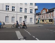 Straßenszene mit drei Personen auf Mopeds an einer belebten Kreuzung, umgeben von mehrstöckigen Fassaden. Im Hintergrund sind ein weißes Gebäude mit Fensterläden und ein rotes Backsteinhaus mit Ladengeschäft erkennbar.