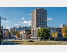 Stadtszene mit einem hohen Wohn- oder Bürogebäude im Zentrum, umgeben von mehrstöckigen Wohnhäusern. Im Vordergrund eine Straße, Grünflächen und ein gelbes Verkehrsschild, dahinter vereinzelt Bäume und parkende Autos.