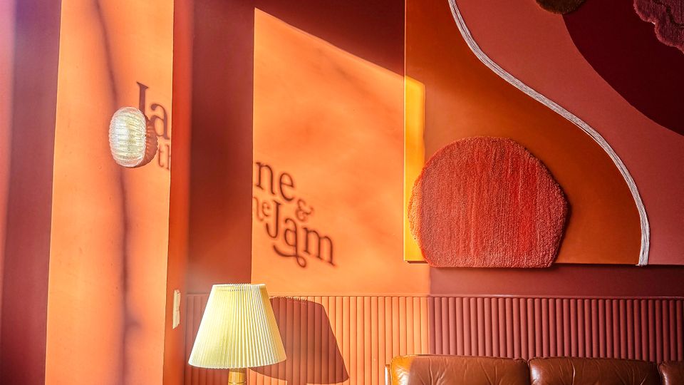 Warm-toned lounge corner with deep coral walls, a brown tufted leather sofa, a pink upholstered chair, and a small marble-topped side table with a pleated lamp. Abstract textured wall art and a round wall sconce are lit by strong sunlight casting long shadows across the wall.
