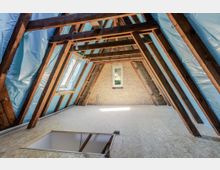 Ausgebauter Dachboden mit sichtbaren Holzbalken, gedämmten Dachschrägen mit blauer Folie und hellem Holzplattenboden. An der Stirnseite sind eine Ziegelwand sowie ein kleines Fenster und ein Dachfenster an der linken Schräge zu sehen.