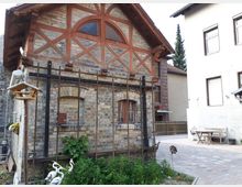 Kleines Backsteingebäude mit Fachwerk-Giebel und halbrundem Fenster im oberen Bereich, davor ein Rankgerüst an der Fassade. Daneben liegt ein gepflasterter Innenhof mit Sitzbank und Tisch zwischen angrenzenden Wohnhäusern.