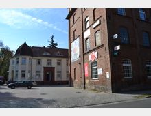 Zweistöckiges Gebäude mit hellem Putz und rotem Ziegeldach links, daneben ein rotes Backsteingebäude mit großen Bogenfenstern und Werbeschildern. Ein gepflasterter Platz und ein geparkter Pkw befinden sich im Vordergrund.