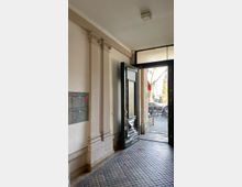 Eingangsbereich eines Altbaus mit hellen Wänden und dezenten Stuckverzierungen an den Wandpfeilern. Links befinden sich Briefkästen, während rechts eine geöffnete, schwarze Tür mit Glaseinsatz ins Freie führt, wo Fahrräder und Teile einer Straße erkennbar sind.