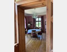 Ein Esszimmer mit Holzboden und einer ovalen Holztafel, die mit einer weißen Spitzendecke bedeckt ist, umgeben von Stühlen mit gepolsterten Sitzflächen. An der Decke hängt ein dekorativer Kronleuchter, und ein großes Fenster mit Vorhängen sorgt für Tageslicht.