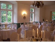 Ein festlich dekorierter Veranstaltungsraum mit weiß gedeckten Tischen und Stühlen, die mit Hussen bezogen sind. Der Raum hat große Fenster mit Ausblick ins Grüne, Wandleuchter und einen zentralen Kronleuchter sorgen für Beleuchtung, während Kerzen auf den Tischen eine elegante Atmosphäre schaffen.