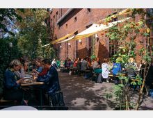 Außenbereich eines Cafés mit Holzdielenboden, zahlreichen Sitzbänken und Tischen, an denen Menschen essen und sich unterhalten. Im Hintergrund eine rote Backsteinfassade mit hohen Fenstern, darüber gelbe Markisen; umgeben von Bäumen und viel Grün.