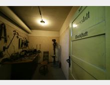 Ein kleiner Werkstattraum mit hellgelber Tür, auf der "Werkstatt" steht. Im Inneren befinden sich Werkzeuge und Geräte auf einer Arbeitsfläche an der linken Wand, eine Standbohrmaschine in der Ecke und eine einfache Deckenbeleuchtung.