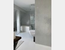 Modernes Badezimmer mit weißen Bodenfliesen und Wänden, die mit kleinen, schimmernden Mosaikfliesen verkleidet sind. Zu sehen sind eine wandmontierte Toilette, eine offene, verglaste Dusche und eine freistehende Badewanne.