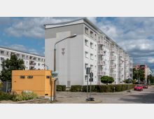 Eine Wohnsiedlung mit mehrstöckigen weißen Wohnblöcken, die über Balkone an der Fassade verfügen. Im Vordergrund steht ein gelber Imbisscontainer mit der Aufschrift "Döner Kebap", daneben Gehwege, ein Baum und ein Parkplatz mit geparkten Autos.